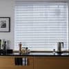 White horizontal Venetian blinds covering a large window, slats mostly closed and filtering diffuse daylight; modern kitchen countertop beneath holds jars, a kettle, utensil holder and wooden cabinets, bright interior.