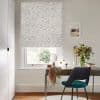 A patterned Roman blind (white with pale leaves), fully lowered and filtering daylight; over a window above a desk with mirror, vase of flowers and teal chair in a study.