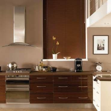 Dark brown horizontal venetian blinds covering a window, fully closed and filtering warm light; modern kitchen with dark wood cabinets, stainless-steel range and hood, countertop appliances, potted orchid on sill.