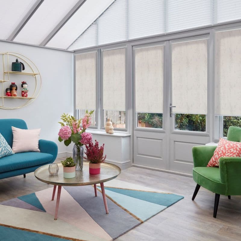 Cream patterned roller blinds cover tall glazed doors and sidelights, partially lowered and filtering soft daylight; bright conservatory-style living room with teal sofa, green armchair, pastel geometric rug, coffee table.