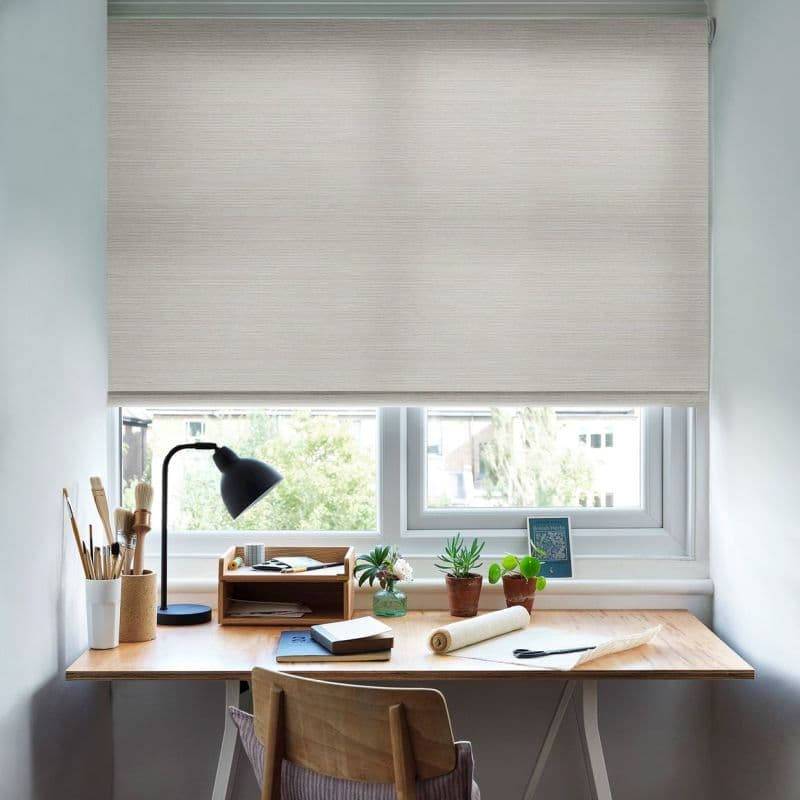 Beige fabric roller blind, lowered almost fully, filtering soft daylight; above a window over a wooden desk with lamp, potted plants, brushes and notebooks in a bright home workspace.