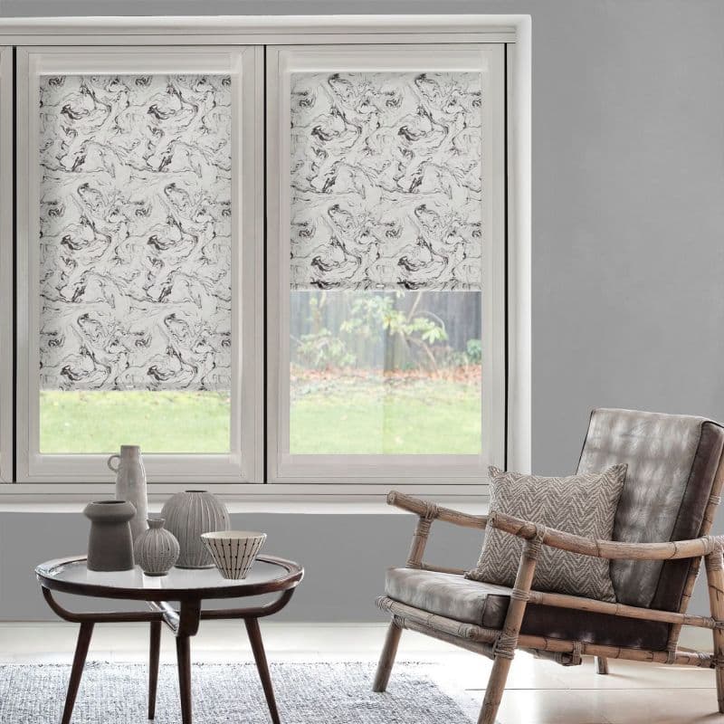 Marble-pattern roller blinds, lowered halfway on triple-pane windows, partially blocking and diffusing daylight; framed by white casings in a grey living room with a rattan armchair and coffee table.