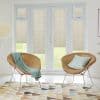 Beige horizontal-slat blinds fitted inside French doors and sidelights, lowered and closed, filtering soft daylight; modern living area with two rattan chairs, pastel cushions, patterned rug and lawn view.