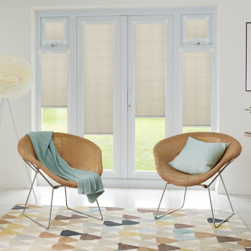 Beige horizontal-slat blinds fitted inside French doors and sidelights, lowered and closed, filtering soft daylight; modern living area with two rattan chairs, pastel cushions, patterned rug and lawn view.