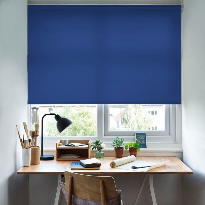 Blue roller blind, lowered across a double window, solid deep-blue fabric blocking most daylight; a sunlit home desk below with lamp, art supplies, potted succulents and an outside view.
British Herbs