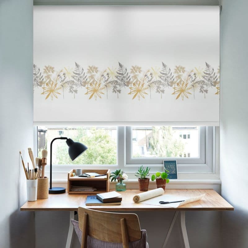 A white roller blind with a horizontal band of yellow-gray botanical prints, drawn down and filtering soft daylight over a small home workspace with desk, lamp, plants, brushes.