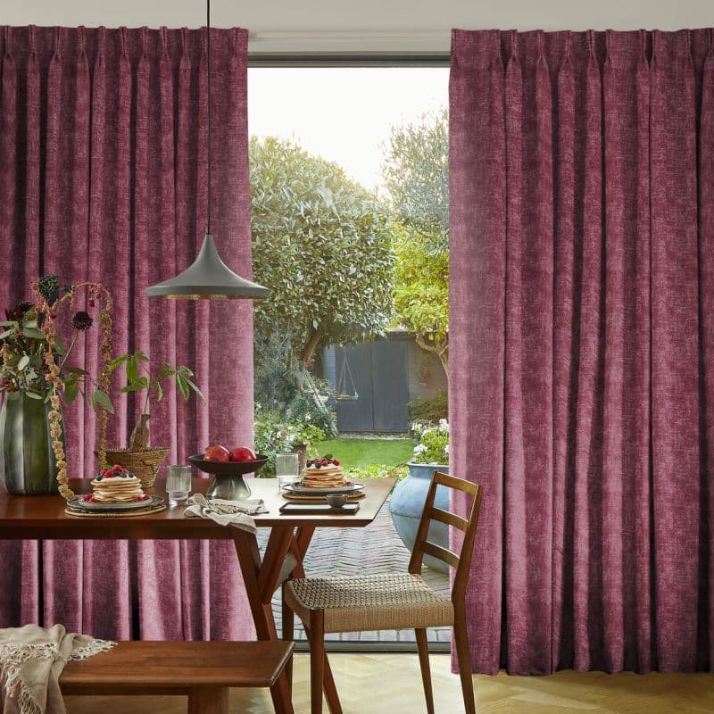 Full-length pleated velvet curtains, deep mauve, drawn open slightly at center to frame and filter daylight through a sliding glass door; dining table with pendant lamp, plates of pancakes, garden beyond.