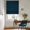 Dark teal Roman blind, lowered with soft horizontal folds, filtering daylight over a white window in a bright home office; wooden desk with vase of flowers and a teal chair.