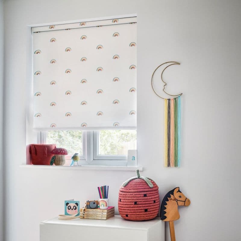 White roller blind printed with small rainbows lowered over the window, filtering soft daylight; nursery shelf and dresser display toys, a strawberry basket and crescent wall-hanging with pastel tassels.
a
