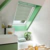 Pleated skylight blind (white), partially lowered over a roof window, filtering daylight. In an attic bathroom with mint-green window frame, white herringbone tiles, sink, bathtub, plant and toiletries.