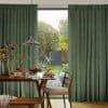 Floor-to-ceiling green textured pleated curtains hang in soft folds, slightly parted to reveal a sunlit garden; a dining table set for breakfast with pancakes, pendant lamp and wooden chairs.