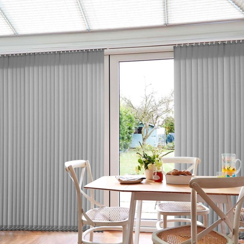 Vertical grey fabric blinds, floor-to-ceiling and mostly closed with a central gap; parted to reveal a glass patio door. Small dining table with chairs, vase, pastries, and daylight-filled garden view.