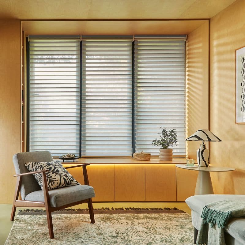 Layered horizontal roller shades across a three-panel window, evenly lowered with alternating sheer and opaque stripes, filtering soft daylight into a warm wood‑paneled sitting alcove with armchair and potted plant.
