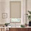 Beige patterned roller blind lowers over a kitchen window, filtering light; stainless faucet and sink sit below on a countertop with bowls of leafy greens, potted plants, and a framed sketch.