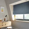 Blue Roman Roman blind lowered in layered folds, covering the upper casement window and partially filtering daylight; small sloped‑ceiling home office with light wood desk, lamp, stool, vase on sill. 02 JUNE