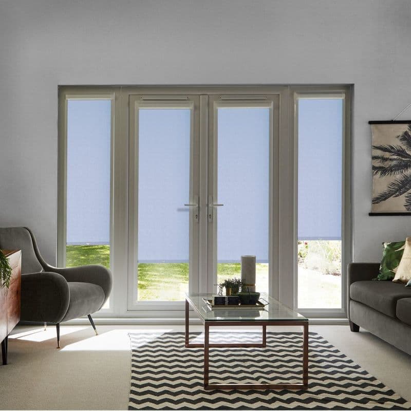 Light-blue roller blinds cover four tall glass panels, lowered with narrow bottom gaps; filtering daylight; living room with gray armchair and sofa, coffee table on rug, lawn visible outside.