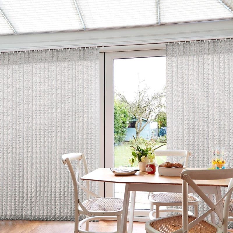 Patterned vertical blinds on a top rail, mostly closed with a central gap; filtering soft daylight while revealing a bright backyard; over a sunlit dining nook with table and chairs.