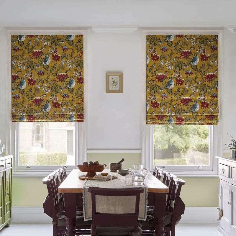 Two mustard-yellow floral Roman blinds, lowered over adjacent windows, patterned with red and blue flowers, filtering daylight into a dining room with wooden table, chairs and garden view.