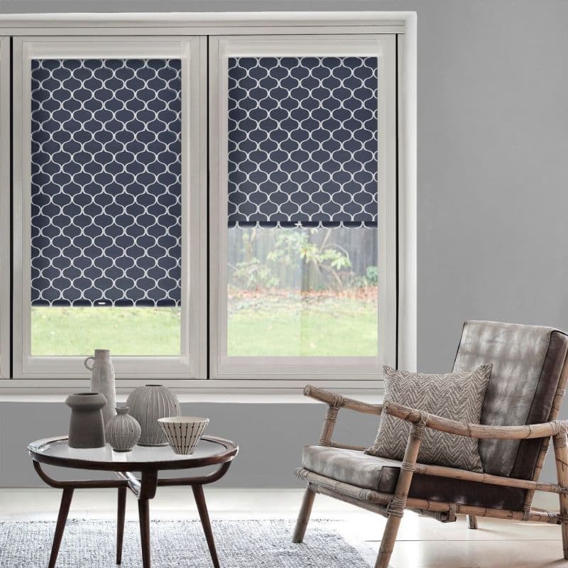 Navy-blue roller blinds with white trellis pattern, left fully lowered, right half-lowered, blocking light; set in a gray-walled living room with a rattan armchair and small table.