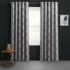 Grommet-top silver-gray curtains with dark geometric lattice, parted at center over a window, filtering soft daylight; dark charcoal walls, small wooden side table with glass-dome lamp, framed abstract art.