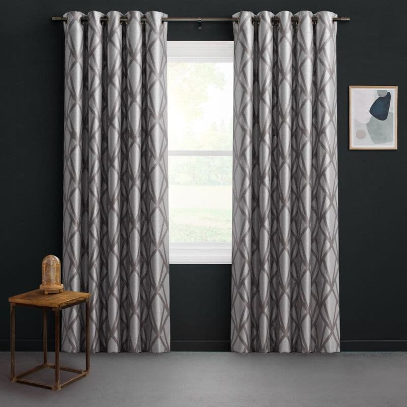 Grommet-top silver-gray curtains with dark geometric lattice, parted at center over a window, filtering soft daylight; dark charcoal walls, small wooden side table with glass-dome lamp, framed abstract art.