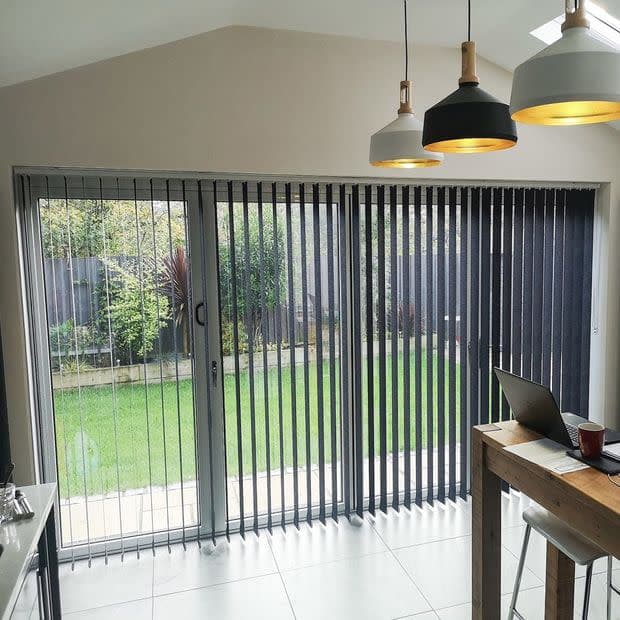 Vertical dark grey vertical blinds covering sliding glass patio doors, slats angled to filter light and reveal a garden; kitchen/dining interior with pendant lights, tiled floor, wooden table and laptop.
