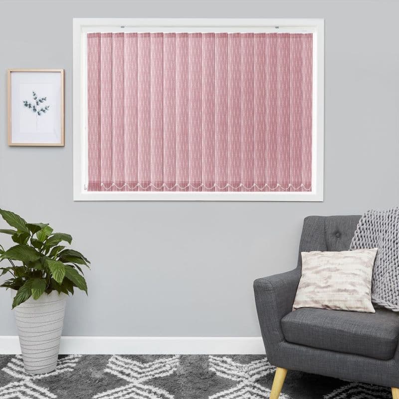 Pink textured vertical blinds cover a white-framed window, fully closed and softly filtering daylight; in a gray living room with potted plant, framed botanical print, gray armchair and patterned rug.