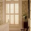 White plantation shutters covering a tall arched window, closed and filtering warm daylight; positioned on a stair landing with floral wallpaper, a wooden banister, side table and vase of lilies.