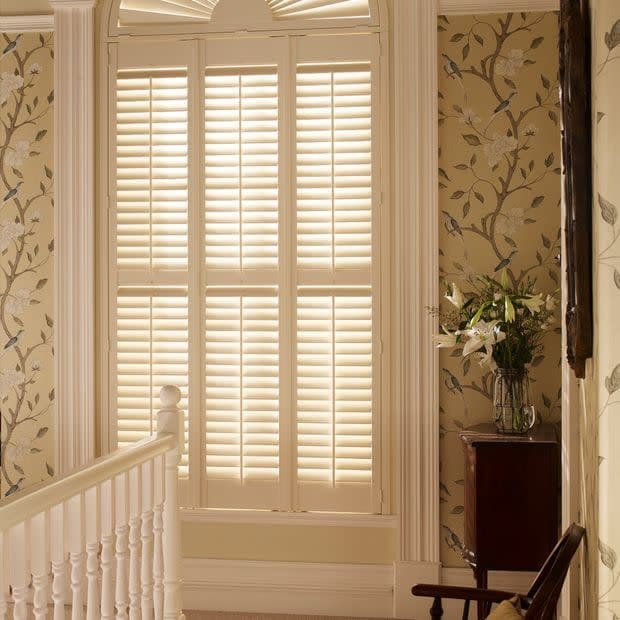White plantation shutters covering a tall arched window, closed and filtering warm daylight; positioned on a stair landing with floral wallpaper, a wooden banister, side table and vase of lilies.
