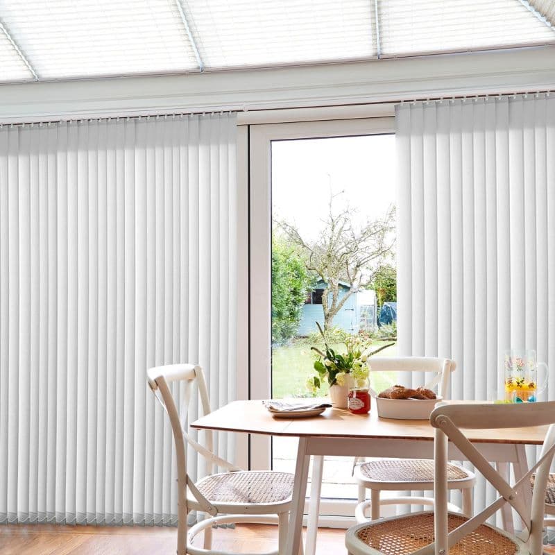 White vertical blinds, mostly closed but parted at center, filtering daylight and revealing a patio door; small dining table with wooden chairs, vase of flowers, and garden beyond.