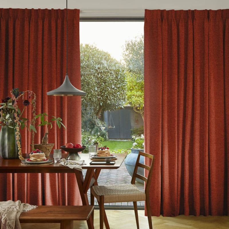 Heavy rust-red pleated curtains, pulled to each side and parted to reveal a glass door, filtering soft daylight over a dining table set for breakfast and a green backyard.