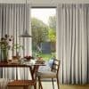 Beige floor-length pleated curtains, drawn partly aside to softly filter daylight through a sliding door; a wooden dining table with pancakes inside opens onto a sunlit garden path and shed.