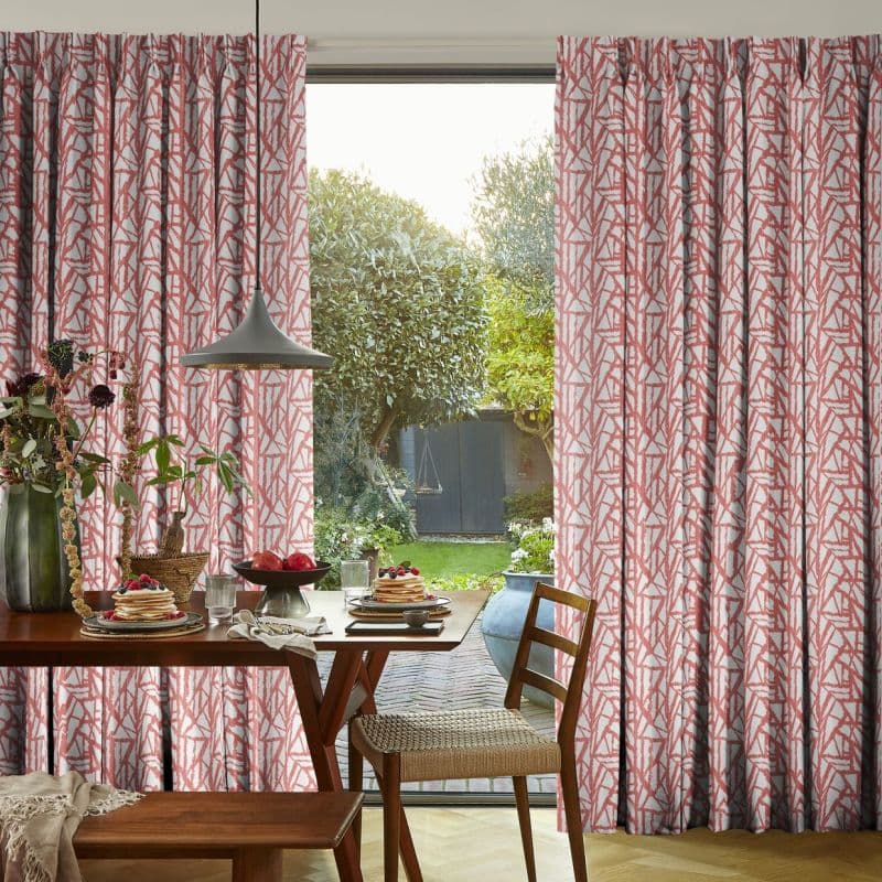 Pleated coral-and-white patterned curtains, parted centrally to expose a sliding glass door; framing a sunlit dining nook with breakfast table and leafy backyard trees and a small shed.