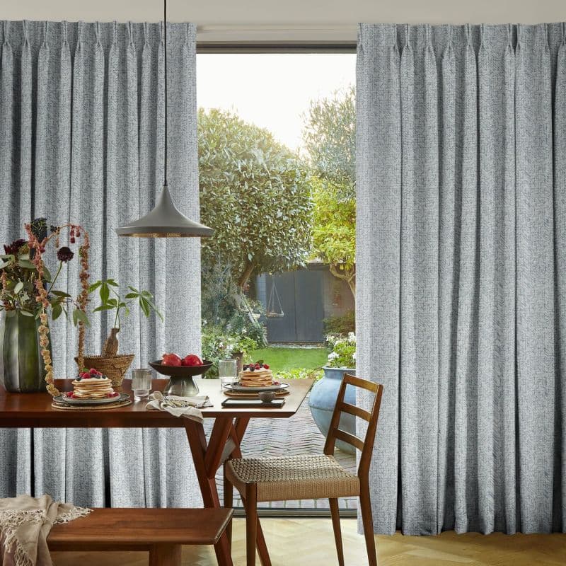 Pleated grey full-length curtains, drawn aside to frame the window opening and filter daylight, in a dining room with a set table, pendant lamp and a sunlit garden visible beyond.