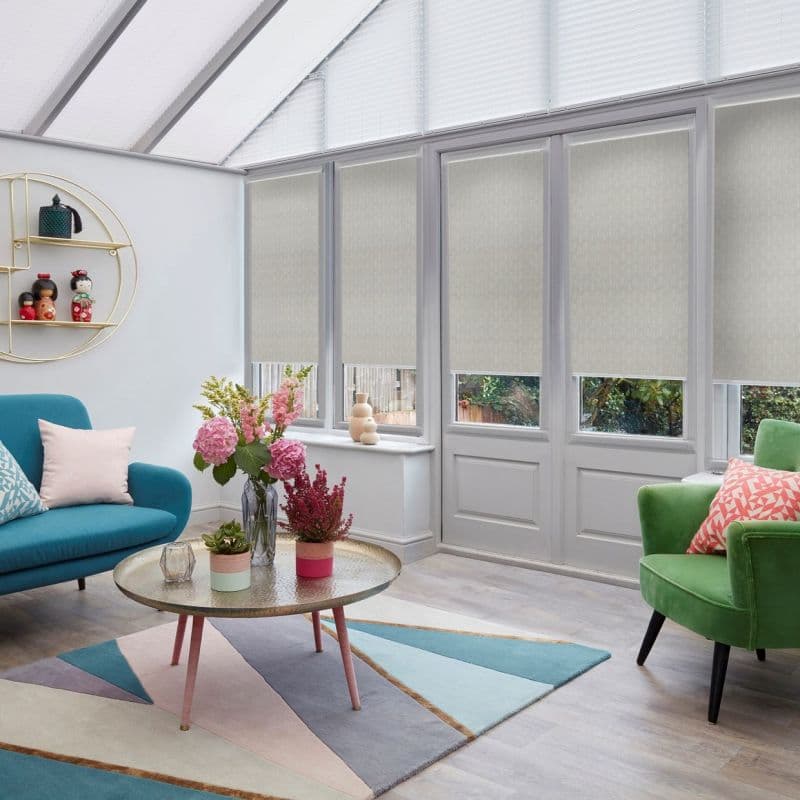 Pale patterned roller blinds lowered over tall conservatory windows, blocking most light with small lower openings; bright sunroom furnished with teal sofa, green armchair, round coffee table, floral arrangement.
