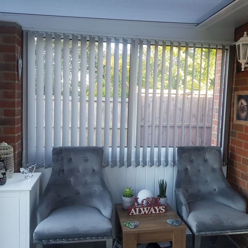 Vertical white blinds cover a wide conservatory window, slats slightly angled and filtering daylight. Two grey tufted chairs flank a wooden side table amid brick walls and a fenced garden view.
LOVE ALWAYS