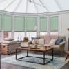Mint-green cellular blinds lowered halfway across white-framed conservatory windows, filtering soft daylight; cozy sunroom living area with beige sofa, chunky pink throw, wooden coffee table and blue rug.