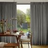 Floor-length grey pleated curtains, drawn open at the center and hanging in neat folds; framing a sunlit garden view, with a timber dining table set for breakfast and a hanging pendant lamp.