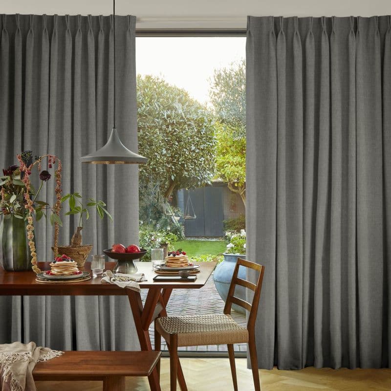 Floor-length grey pleated curtains, drawn open at the center and hanging in neat folds; framing a sunlit garden view, with a timber dining table set for breakfast and a hanging pendant lamp.