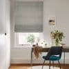 Grey fabric Roman blind, lowered almost to the sill with folded tiers and textured finish; filtering daylight through a narrow bottom gap; above a desk with teal chair, small mirror, vase of flowers, neutral walls.