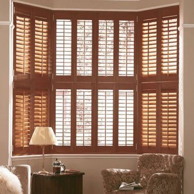 Bay window covered by wooden plantation shutters—multi-panel, upper and lower sections with tilted slats; filtering warm daylight into a living room with upholstered armchairs, lamp, and side table.