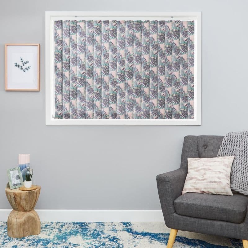 Vertical patterned blinds covering large window, pale pink with teal and gray leaf print; closed, diffusing light; modern living room with gray armchair, wooden side table, framed art, blue rug.