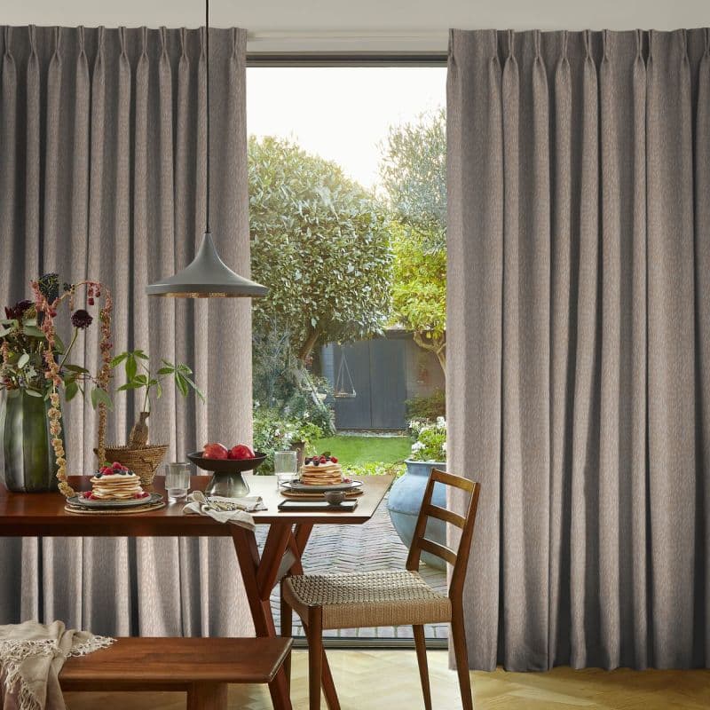 Floor-length gray pleated curtains with pinch pleats, pulled aside to frame a glass door and admit daylight; wooden dining table set for breakfast, pendant light, garden and shed beyond.