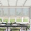 Dark wooden dining table set with six white plates, green glass candleholders and small candles, surrounded by green upholstered chairs; sunlit conservatory with translucent roller blinds and grassy yard beyond.