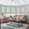 Light-green pleated cellular blinds, lowered halfway across tall white-framed conservatory windows, gently filtering soft daylight; a cozy living area with beige sofa, wooden coffee table, textured rug and plants.