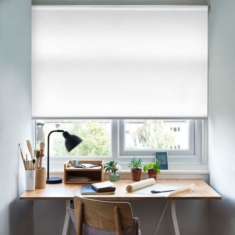 A white roller blind, nearly fully lowered over a twin window, diffuses daylight; a wooden desk below holds a lamp, brushes, notebooks and small potted plants in a bright home workspace.