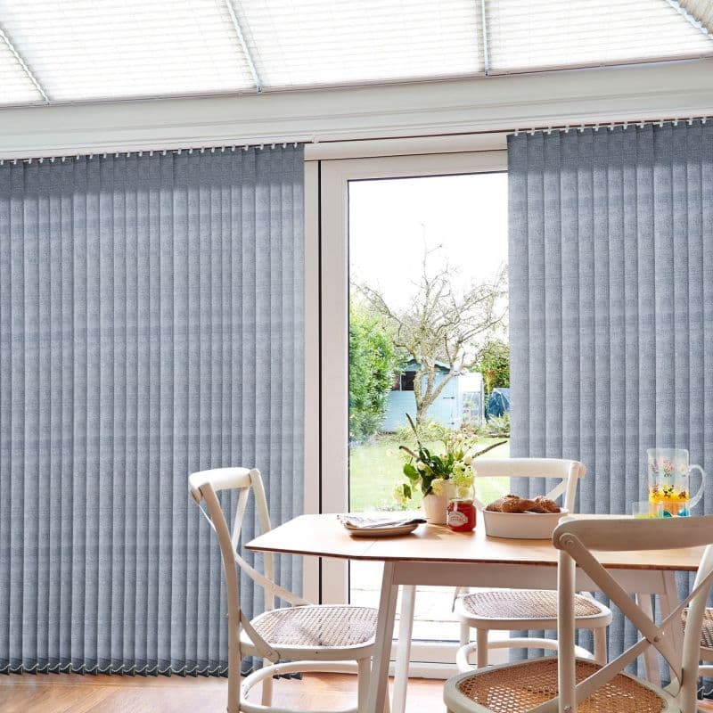Grey vertical fabric blinds covering a sliding glass door, mostly closed but parted centrally to reveal a garden; filtering soft daylight over a wooden dining table with chairs.