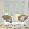 White pleated blinds cover French doors and sidelights, lowered and filtering daylight with lower edges raised to reveal lawn; in a bright room with two wicker chairs and patterned rug.