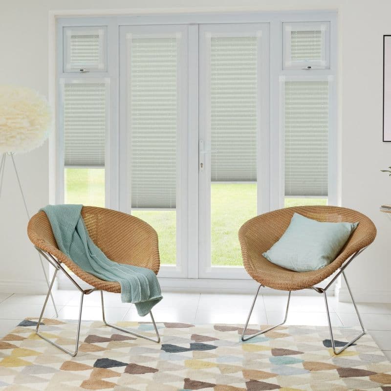 White pleated blinds cover French doors and sidelights, lowered and filtering daylight with lower edges raised to reveal lawn; in a bright room with two wicker chairs and patterned rug.