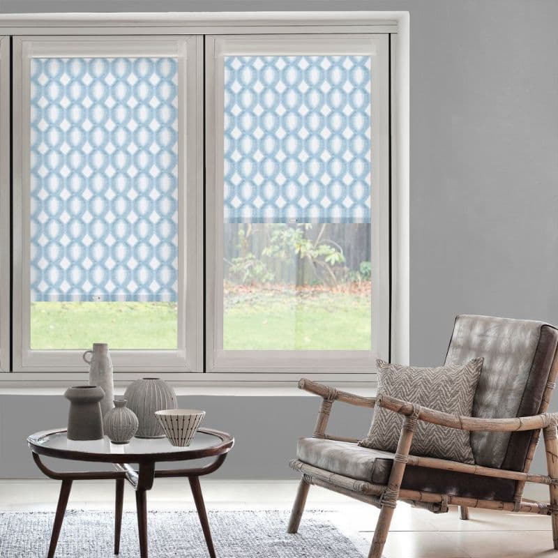 Patterned roller blinds (blue geometric lattice) covering twin windows, one fully lowered and one halfway — filtering daylight and softening neutral living room with rattan armchair, side table, garden view.